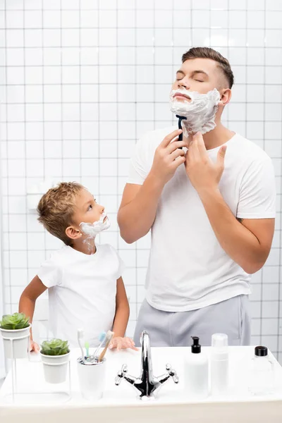 Fils avec mousse à raser sur le visage regardant le père avec les yeux fermés en utilisant un rasoir de sécurité tout en se tenant près de l'évier dans la salle de bain — Photo de stock