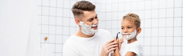 Усміхнений батько дивиться на сина з піною для гоління на обличчі, тримаючи бритву безпеки біля щоки у ванній, банер — стокове фото