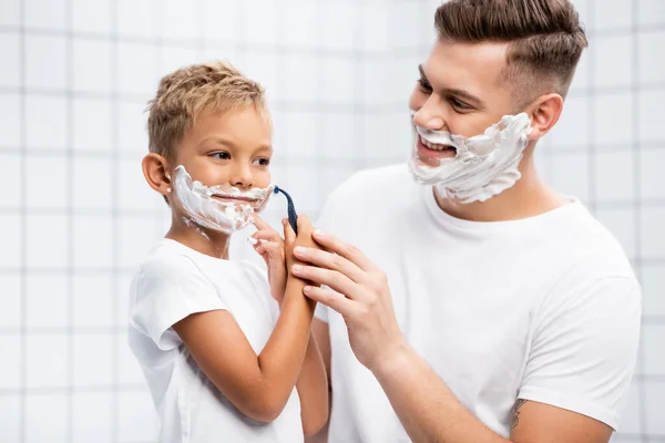Père souriant regardant son fils avec mousse à raser sur le visage tenant rasoir de sécurité près de la joue sur fond flou — Photo de stock