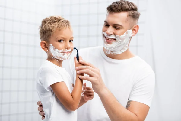 Lächelnder Sohn rasiert sich mit Rasiermesser und blickt in Kamera, während er neben Vater im Badezimmer auf verschwommenem Hintergrund steht — Stockfoto