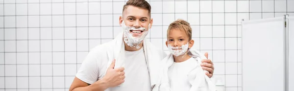 Père et fils souriants avec mousse à raser sur les visages étreignant et regardant la caméra dans la salle de bain, bannière — Photo de stock