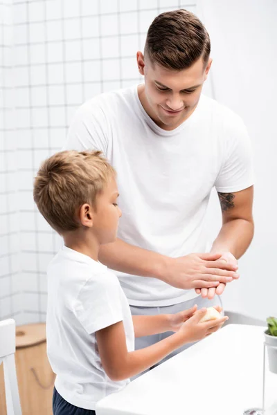 Smiling father washing hands, while looking at son with soap standing near sink in bathroom — Stock Photo