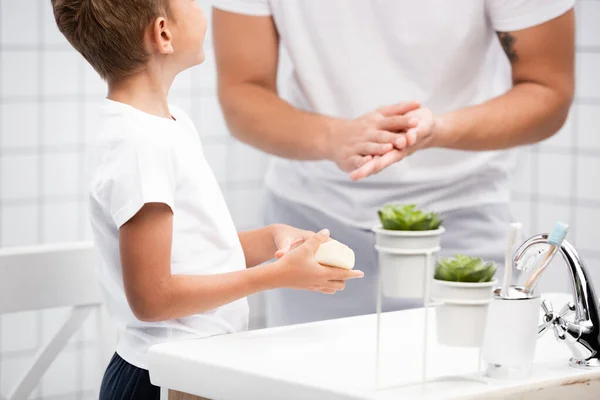 Niño con jabón en las manos de pie cerca de hombre frotándose las manos cerca del lavabo en el baño sobre fondo borroso - foto de stock