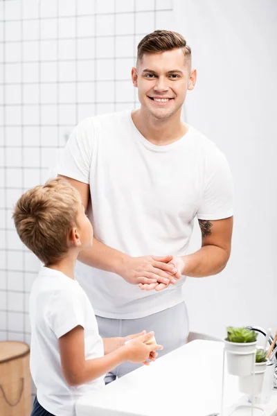 Hijo sosteniendo jabón y de pie cerca sonriente padre frotando las manos mientras mira a la cámara en el baño - foto de stock
