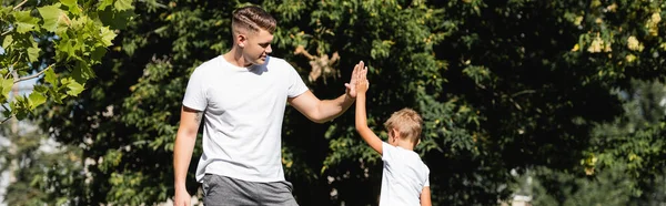 Preschooler son and father in sportswear giving each other high five in park with blurred trees on background, banner — Stock Photo
