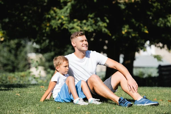 Padre sorridente seduto vicino al figlio in abbigliamento sportivo mentre distoglie lo sguardo nel parco con alberi sfocati sullo sfondo — Foto stock