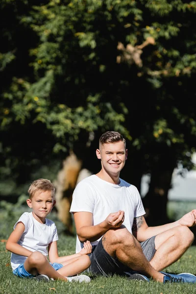 Pai e filho em sportswear olhando para a câmera enquanto relaxa, sentado em pose de lótus na grama no fundo borrado — Fotografia de Stock