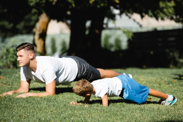 Fils en vêtements de sport faisant planche près du père regardant loin dans le parc sur fond flou — Photo de stock