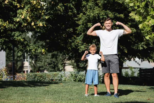 Smiling father and son looking at camera while posing with raised hands showing power in park — Stock Photo