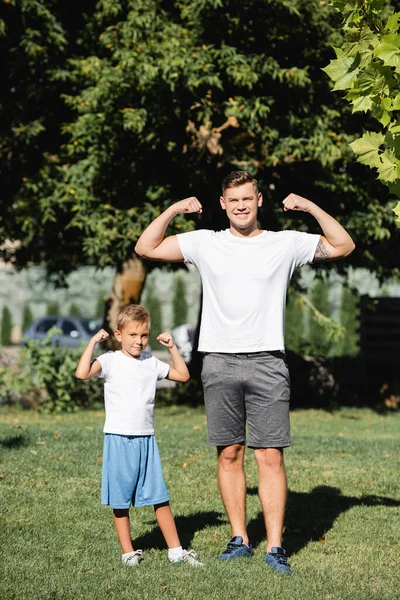 Sonriente padre e hijo en ropa deportiva mirando a la cámara, mientras muestra las manos levantadas con el parque borroso en el fondo - foto de stock