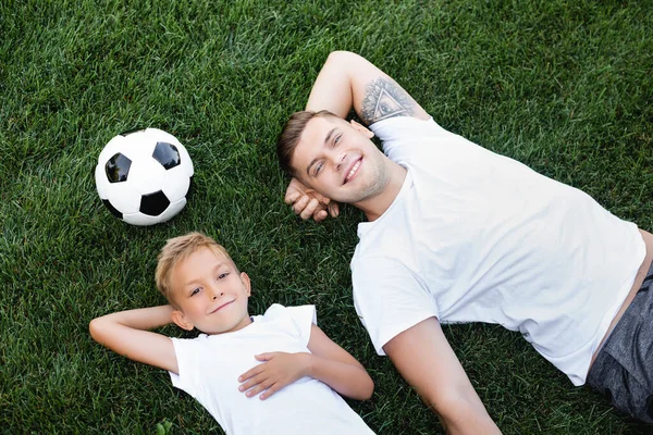 Top view of smiling young adult man and boy with hands behind heads lying near ball on grass — Stock Photo