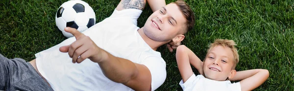 Vue aérienne du fils souriant regardant la caméra tout en étant couché près du père pointant du doigt sur l'herbe, bannière — Photo de stock