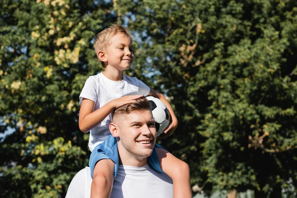 Sonriente hijo con bola a caballo a cuestas en feliz padre en el parque sobre fondo borroso - foto de stock