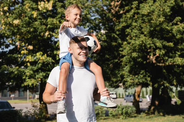 Sonriente hijo sosteniendo la pelota y mostrando el puño, mientras cabalga a cuestas en el feliz padre en el parque sobre fondo borroso - foto de stock