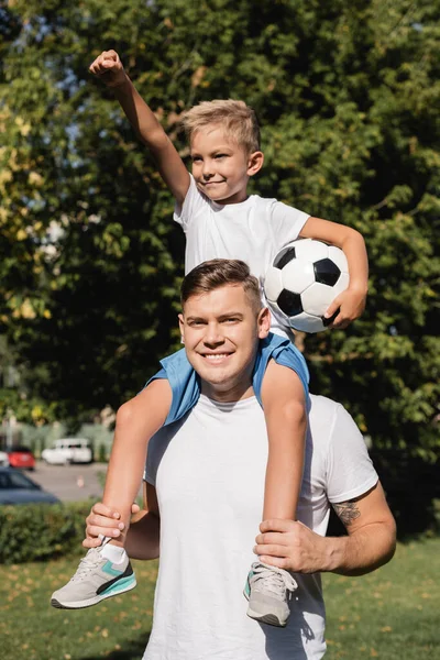 Feliz filho com a mão no ar segurando bola, enquanto montando piggyback no pai sorridente no parque no fundo borrado — Fotografia de Stock