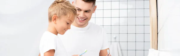 Padre sorridente che guarda il figlio che tiene lo spazzolino in bagno, striscione — Foto stock