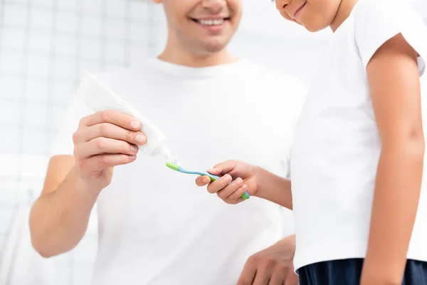 Vista cortada de filho sorridente com escova de dentes em pé perto do pai espremendo pasta de dentes no banheiro no fundo borrado — Fotografia de Stock