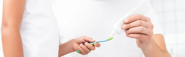 Vista recortada del niño sosteniendo el cepillo de dientes mientras está cerca del hombre apretando la pasta de dientes, pancarta - foto de stock