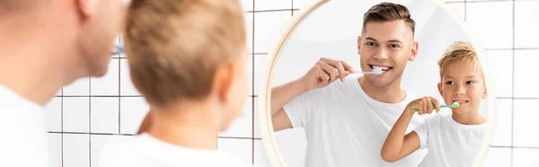 Alegre padre e hijo cepillándose los dientes mientras mira el espejo en el baño en primer plano borroso, pancarta - foto de stock
