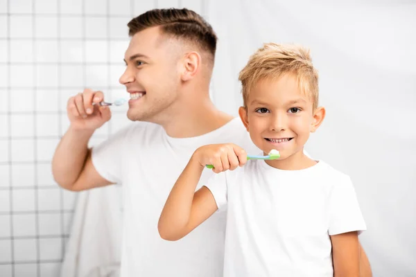 Lächelnder Junge mit Zahnbürste, der Zähne zeigt, während er in die Kamera schaut und neben einem Mann steht, der im Badezimmer wegschaut — Stockfoto