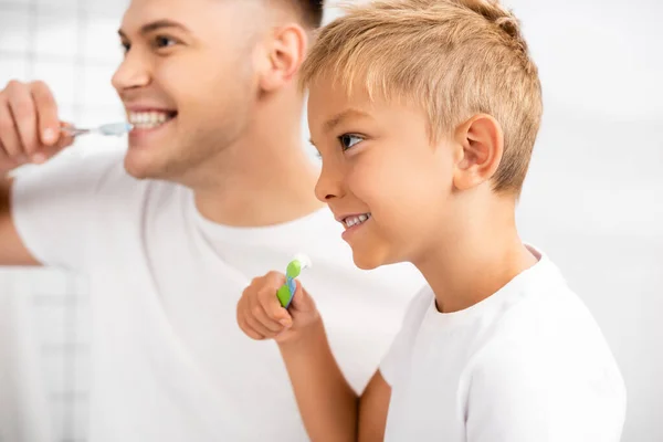 Улыбающийся мальчик с зубной щеткой показывает зубы, глядя в сторону с размытым мужчиной на заднем плане — стоковое фото