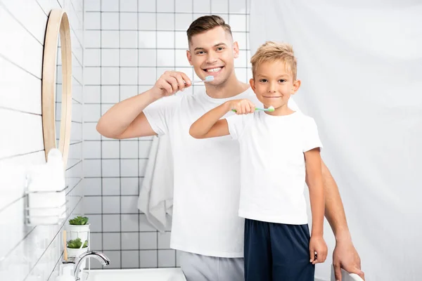 Glücklicher Vater und Sohn mit Zahnbürsten, die in die Kamera schauen, während sie im Badezimmer am Waschbecken stehen — Stockfoto