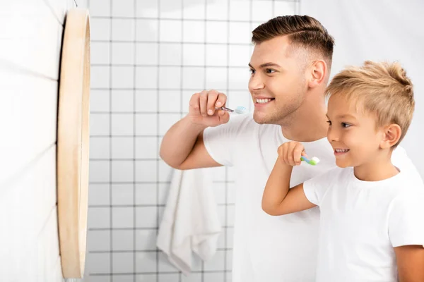 Sonriente padre e hijo sosteniendo cepillos de dientes y mirando al espejo en el baño - foto de stock