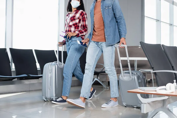 Ausgeschnittene Ansicht einer afrikanisch-amerikanischen Frau in medizinischer Maske mit Pass und Bordkarte, während sie mit einem Mann am Flughafen spazieren geht — Stockfoto
