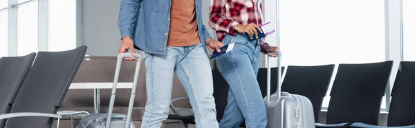 Vista parcial de la mujer afroamericana en máscara médica con pasaporte con tarjeta de embarque mientras camina con el hombre en el aeropuerto, pancarta - foto de stock