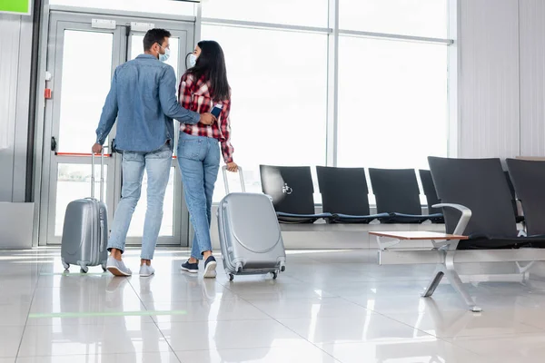 Vista trasera de pareja multicultural caminando con equipaje en el aeropuerto - foto de stock