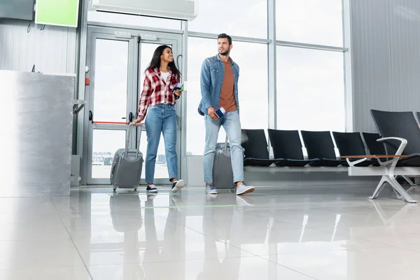 Feliz pareja multicultural caminando con equipaje y pasaportes en el aeropuerto - foto de stock