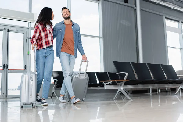 Cheerful multicultural couple walking with luggage in airport — Stock Photo