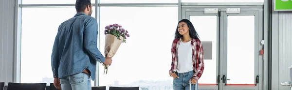 Homem segurando flores embrulhadas ao encontrar feliz namorada afro-americana no aeroporto, bandeira — Fotografia de Stock