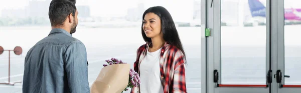 Hombre sosteniendo flores envueltas mientras se encuentra sonriente mujer afroamericana en el aeropuerto, pancarta - foto de stock