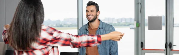 Cheerful man smiling while looking at african american woman with outstretched hands on blurred foreground, banner — Stock Photo
