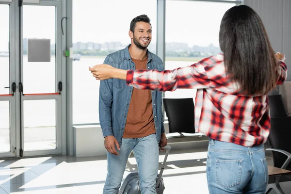 Felice uomo che cammina con i bagagli mentre guarda la donna afro-americana con le mani tese in primo piano sfocate — Foto stock