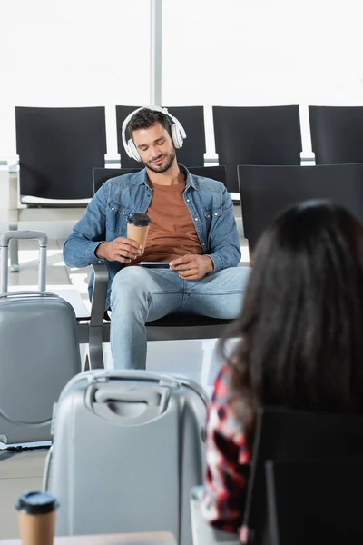 Hombre barbudo con auriculares sosteniendo una taza de papel cerca del equipaje y mujer en primer plano borroso - foto de stock