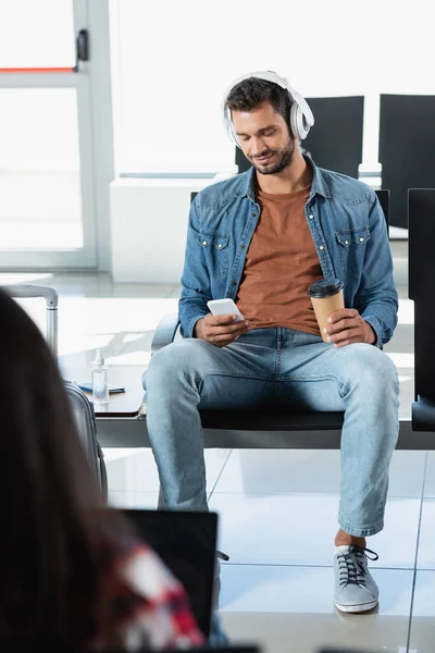 Uomo barbuto in cuffie con tazza di carta e smartphone vicino alla donna in primo piano sfocato — Foto stock