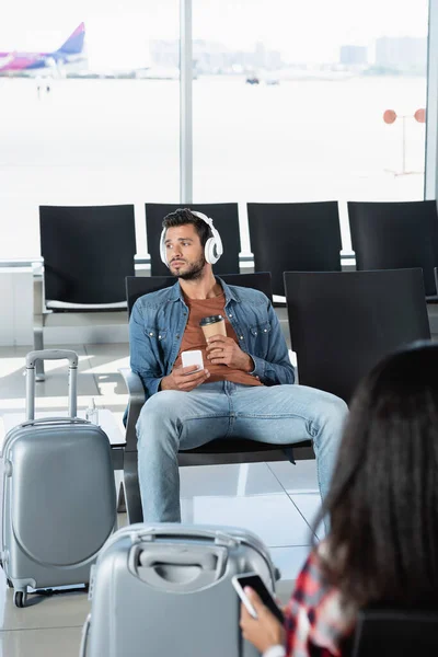 Hombre barbudo en auriculares con taza de papel y teléfono inteligente cerca del equipaje y la mujer en primer plano borrosa - foto de stock