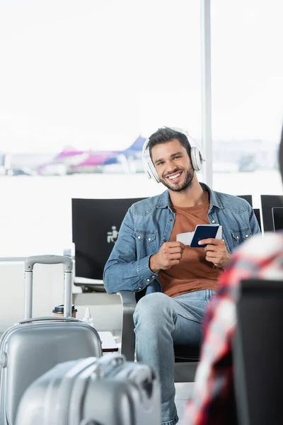Homem feliz em fones de ouvido com passaporte e bilhete aéreo perto do passageiro em primeiro plano turvo — Fotografia de Stock