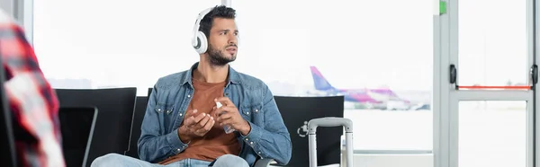Barbudo hombre en auriculares usando desinfectante en el aeropuerto cerca de pasajero en primer plano borrosa, bandera - foto de stock