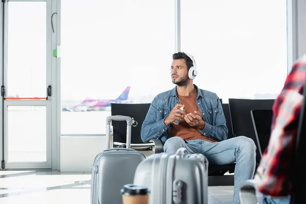 Man in headphones using sanitizer in airport near passenger on blurred foreground — Stock Photo