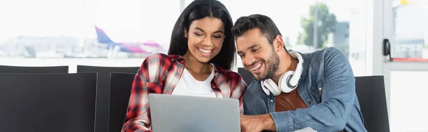Feliz casal interracial olhando para laptop no aeroporto, banner — Fotografia de Stock