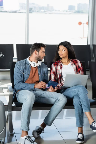 Uomo barbuto in cuffia in possesso di passaporto e guardando la donna afro-americana con computer portatile in aeroporto — Foto stock