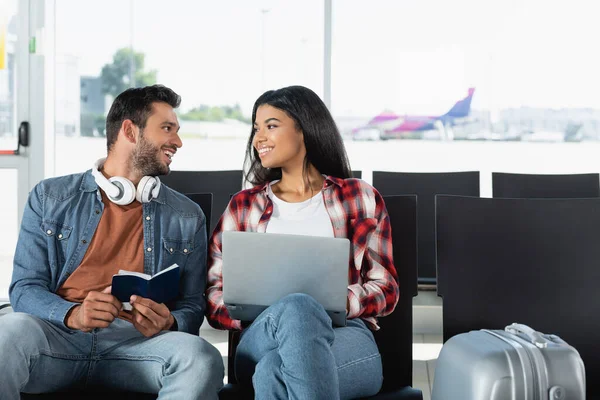 Щаслива міжрасова пара дивиться один на одного біля ноутбука в аеропорту. — стокове фото