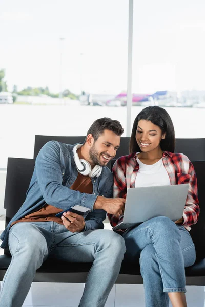 Весела міжрасова пара дивиться на ноутбук в аеропорту — стокове фото