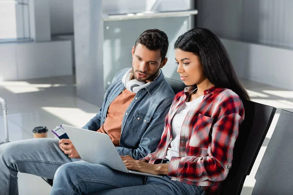 Bearded man in headphones holding passport and looking at african american woman using laptop in airport — Stock Photo