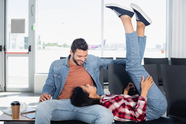 African american woman with raised legs lying near happy man in departure lounge of airport — Stock Photo