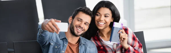 Happy bearded man taking selfie with african american woman holding passport in departure lounge of airport, banner — Stock Photo