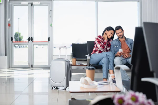Щаслива міжрасова пара дивиться на телефон у відгалуженні аеропорту на розмитому передньому плані. — стокове фото